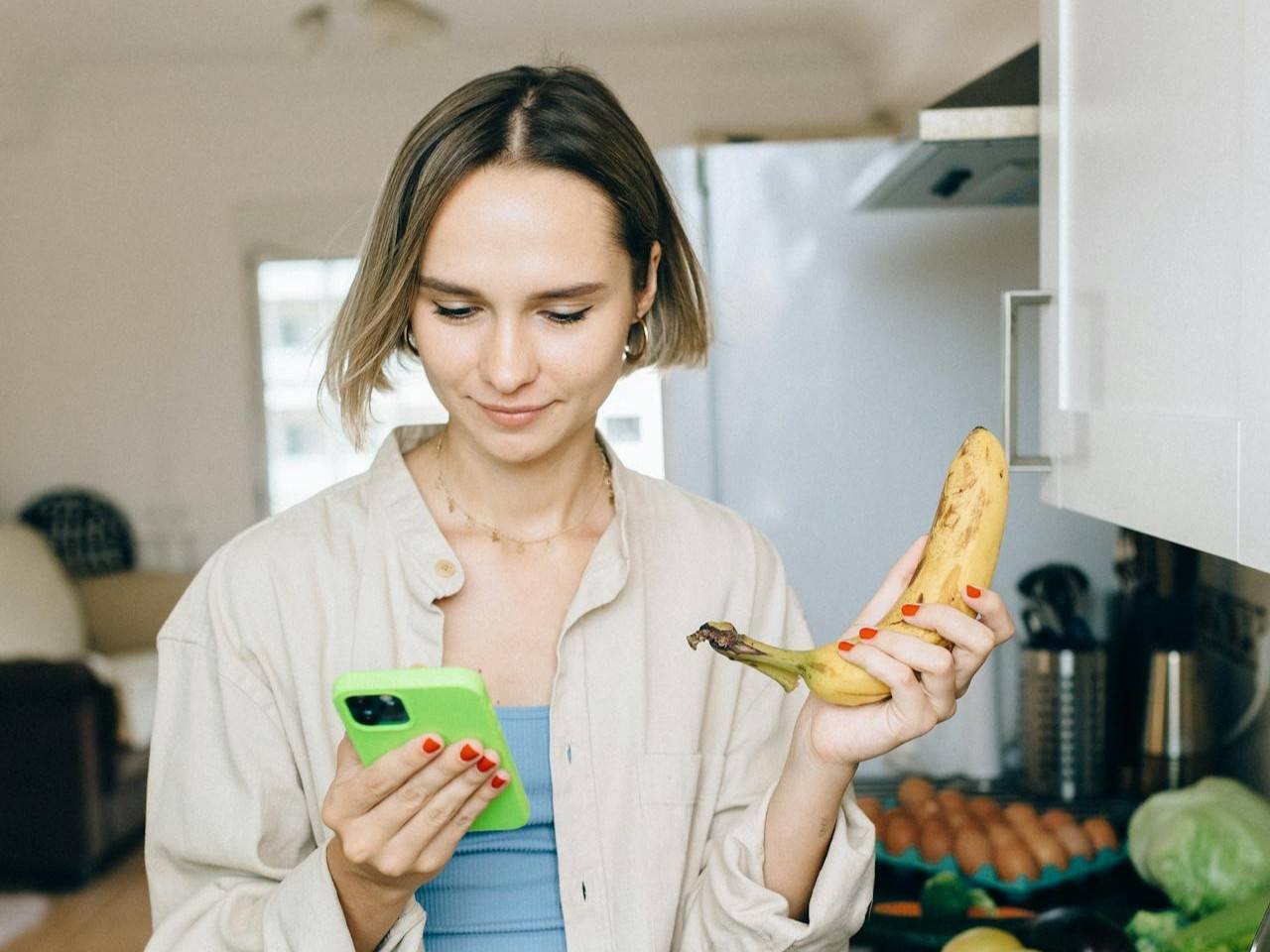 Junge Frau schaut lächelnd auf ihr Smartphone und vergleicht spielerisch verschiedene lange und dicke Penisse. In der Hand hält sie eine Banane als augenzwinkerndes Symbol und wirkt neugierig und amüsiert.